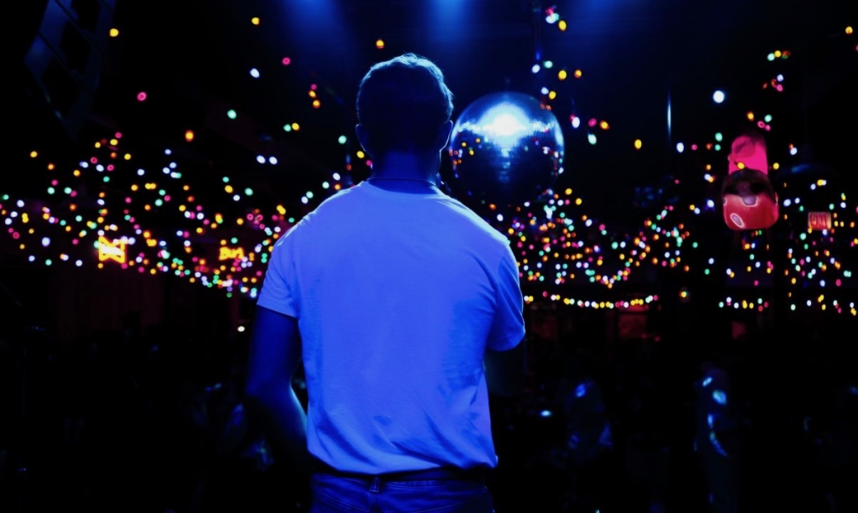Michael Patrick Joyce seen from behind, looking out at a venue full of colorful lights with a disco ball hanging above