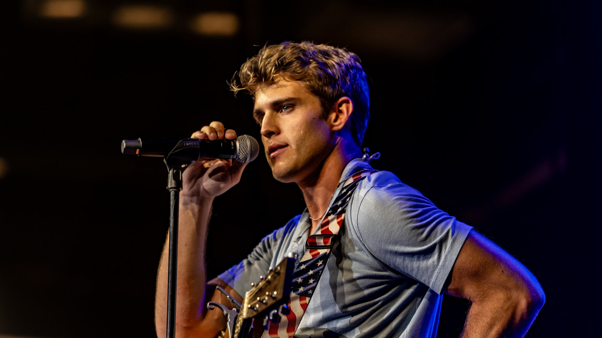 Michael Patrick Joyce singing into a microphone with an American flag guitar strap under blue stage light